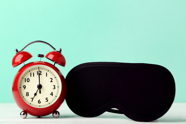 Red old-fashioned alarm clock next to black sleep mask against a turquoise and white background